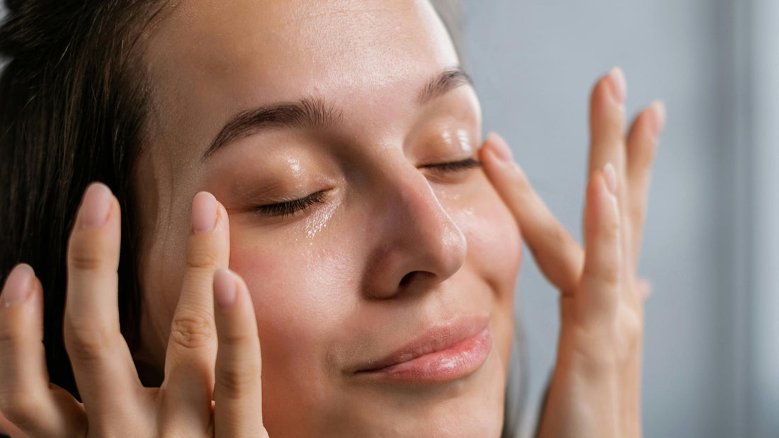 A young woman with clear, glowing skin gently massages a moisturizing serum onto her face with her fingertips, enjoying a self-care skincare routine.