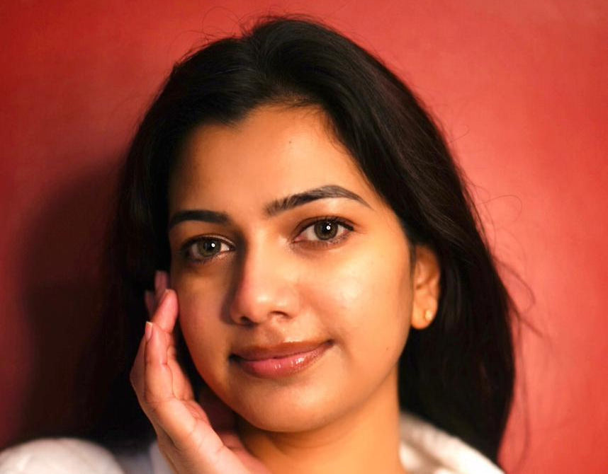 A beautiful Indian woman with radiant, healthy skin applies Donk moisturizer to her cheek. She is wearing a plush white bathrobe and has her hair wrapped in a soft towel, creating a fresh, post-skincare routine look. She smiles gently against a warm red background, exuding confidence and natural beauty. The image highlights the nourishing and hydrating benefits of Donk moisturizer for glowing skin.